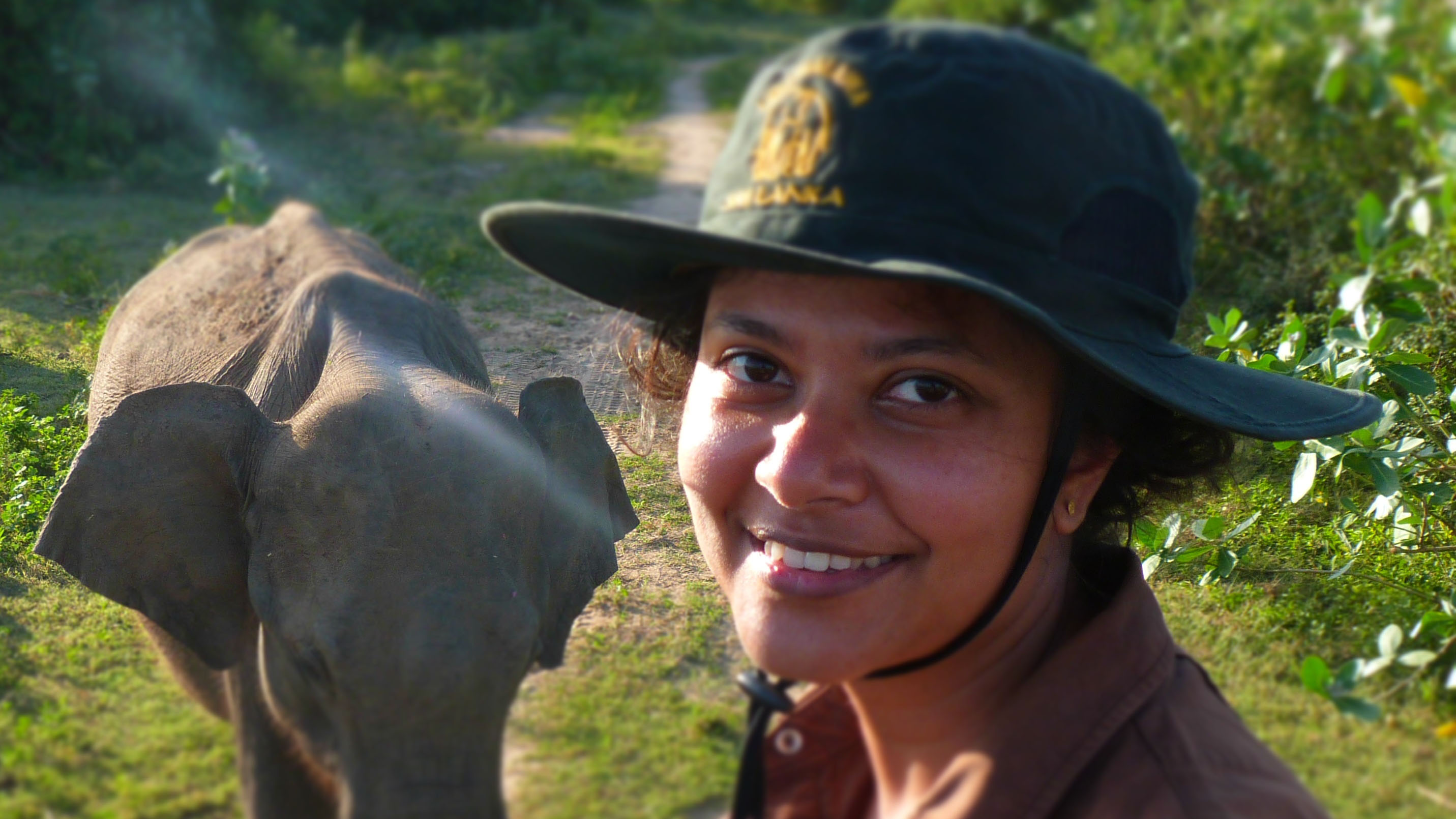 Shermin smiles at the camera, wearing a wide brimmed hat. She is sitting on top of a vehicle, and behind her an Asian elephant is visible.
