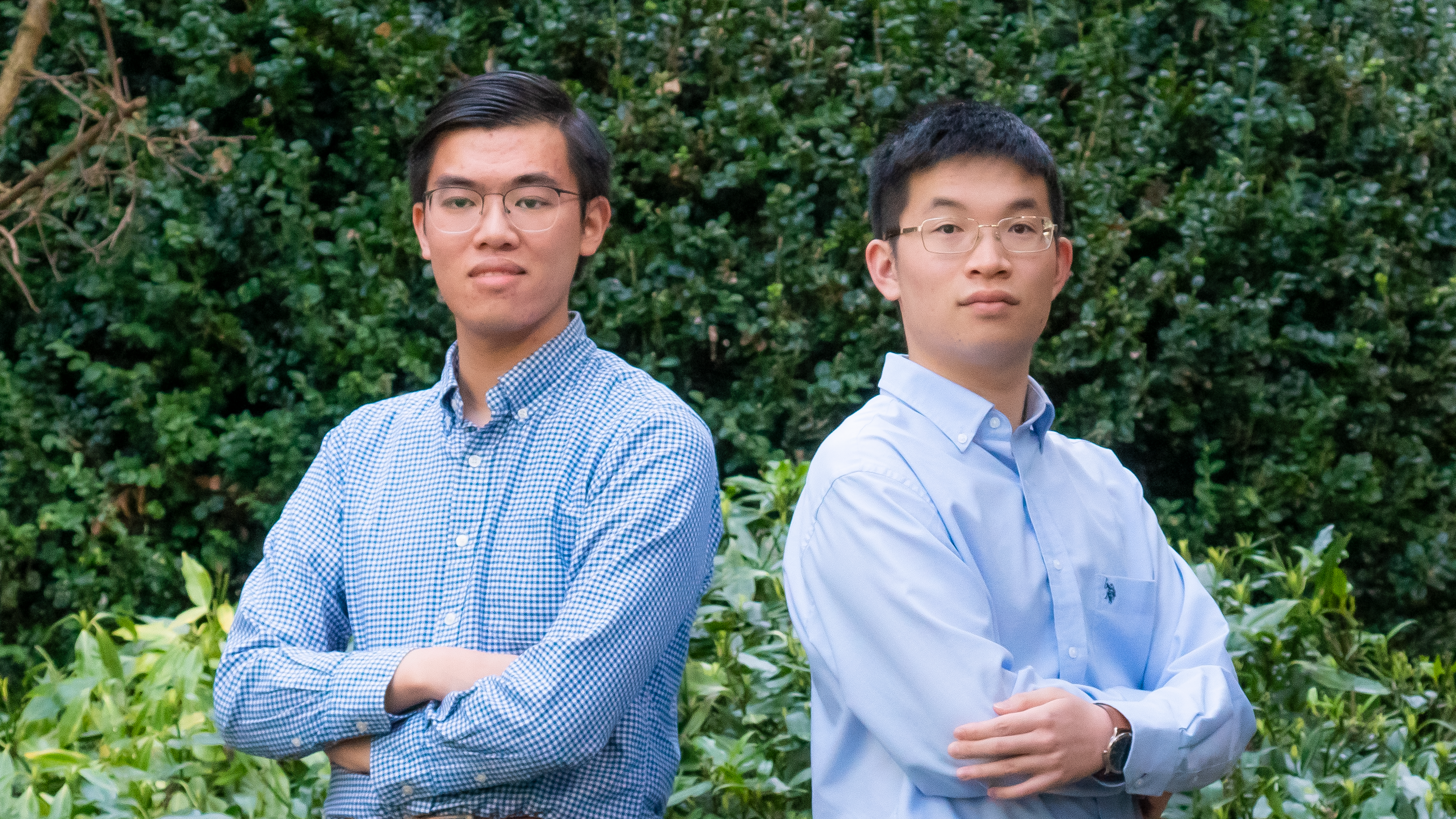 Alvin (left) is a young Asian man wearing a blue short, and Jack (right) is a young Asian man wearing a blue shirt and glasses. Both stand with their arms folded, in front of greenery on campus.
