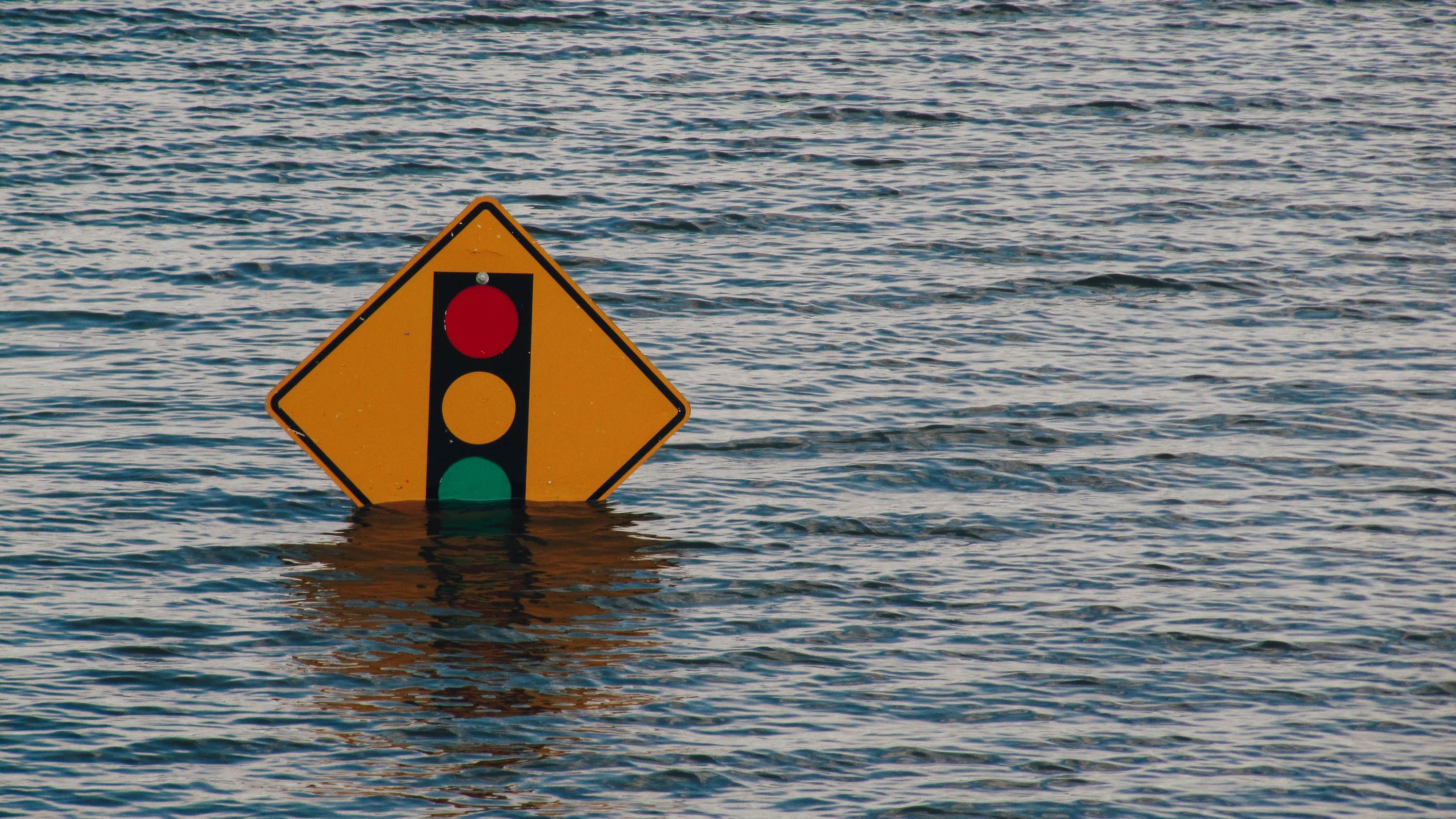 Flood waters partially obscure a road sign depicting a traffic light ahead