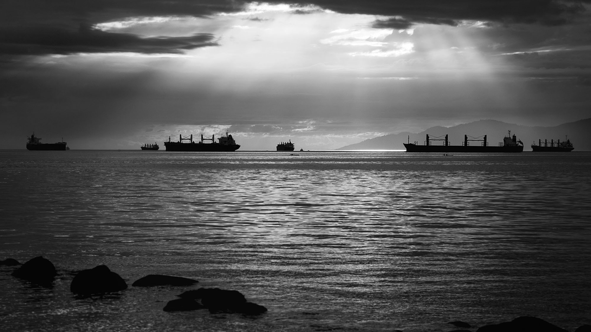 Black and white image of cargo ships in Burrard Inlet. Rays of light shine down from between clouds and Bowen Island is visible in the background.