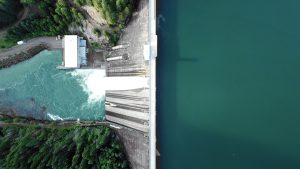 Aerial image: to the right is a large reservoir of dark water, the image is split top to bottom in the middle by the dam itself, and then to the left the spillway and much lower and lighter coloured river are visible, with trees lining the shore.